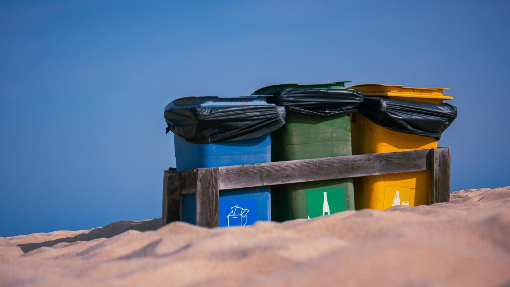Three color-coded outdoor recycling bins placed on sand. 