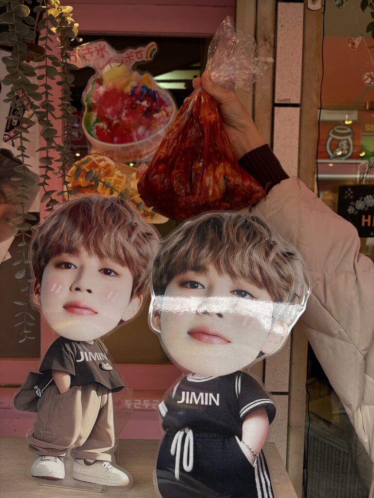 Hand holding a bag of bright red kimchi in front of BTS Jimin standees and a colorful shaved ice sign in Osaka Koreatown