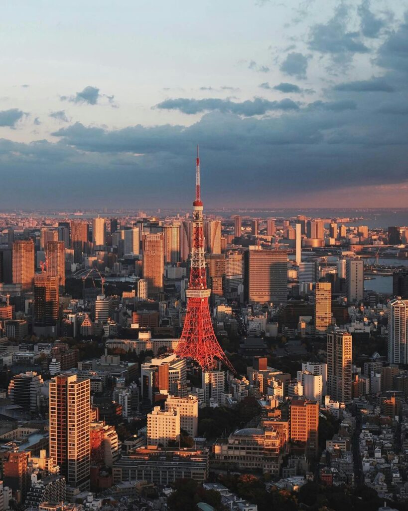 Tokyo Tower rising above the city during dusk, with soft evening light reflecting off surrounding buildings and clouds gathering overhead.