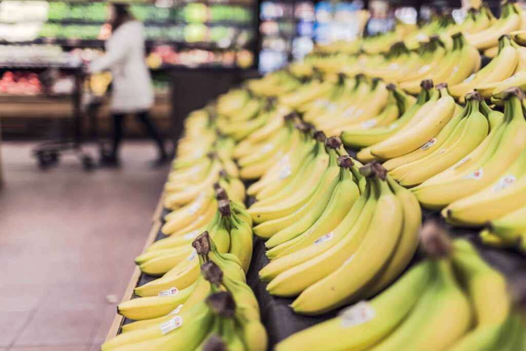 Close-up of rows of bananas displayed in a supermarket, with a shopper and cart blurred in the background.