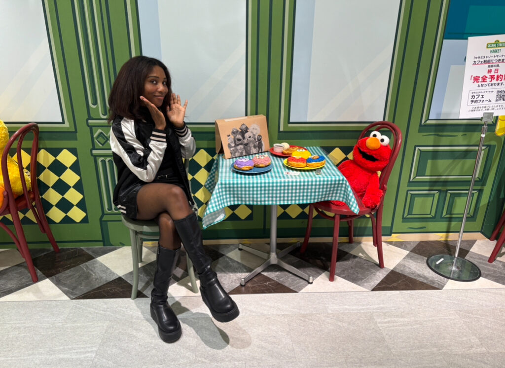A woman posing at a themed café display with Elmo seated at a small table covered in a checkered cloth, surrounded by colorful wall art and toy food.