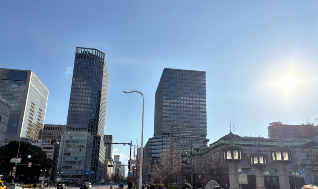 A sunny city street lined with modern office towers and a historic building, with pedestrians walking below a bright blue sky.