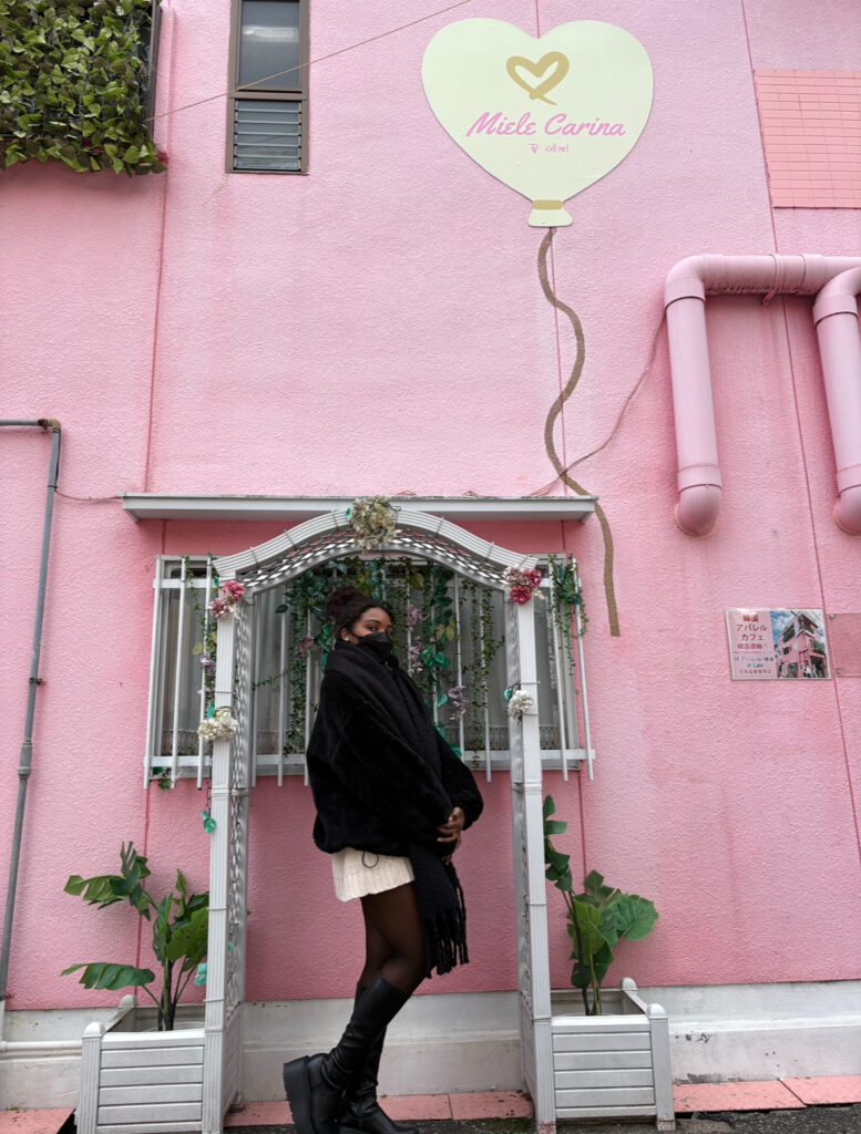 A woman standing in front of a pink café exterior with floral decorations and a heart-shaped sign above the entrance.