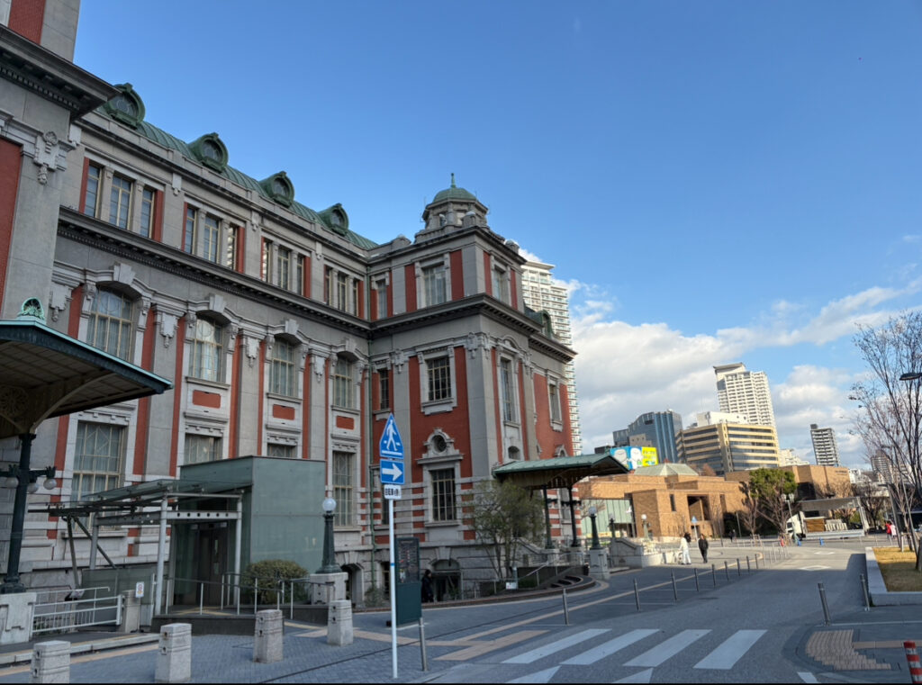 A historic red-brick building in the city framed by a wide street, modern towers, and a clear blue sky.