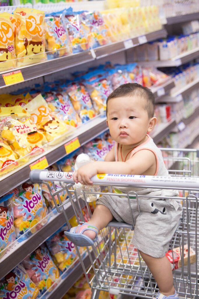 Young child sitting in a shopping cart in a supermarket aisle surrounded by colorful packaged snacks.