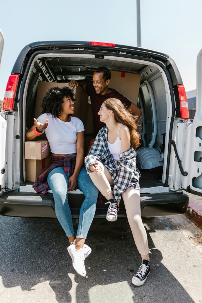 Three people sitting at the back of a moving van surrounded by packed boxes.
