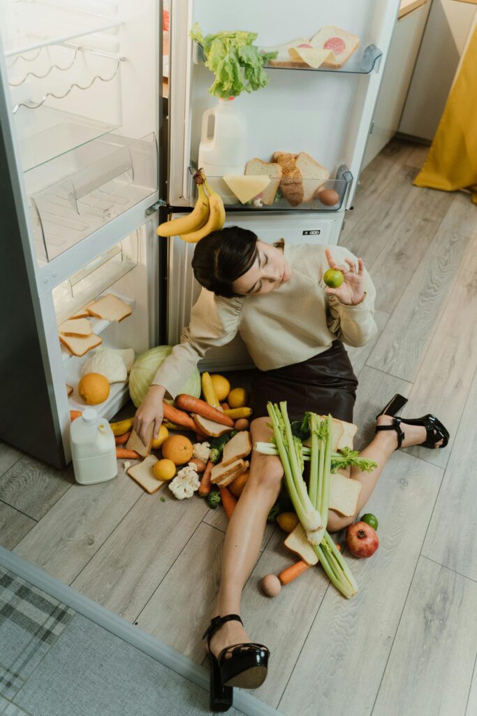 Woman sitting on the floor beside an open refrigerator with food items spread out during packing.