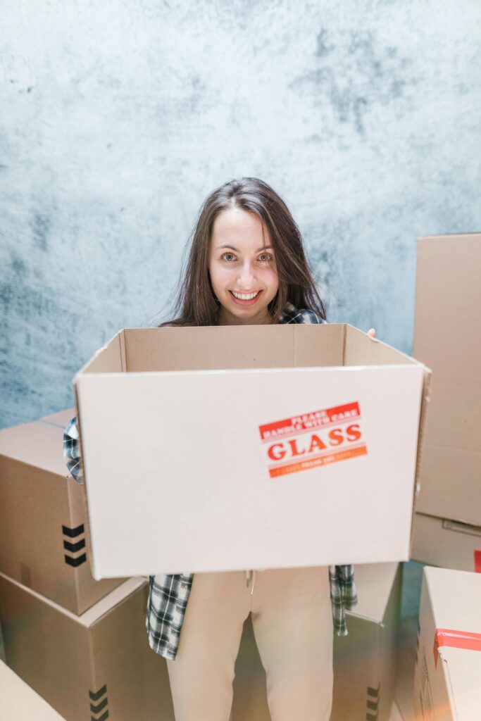 Young child peeking out from inside a large cardboard moving box while family members unpack in the background.