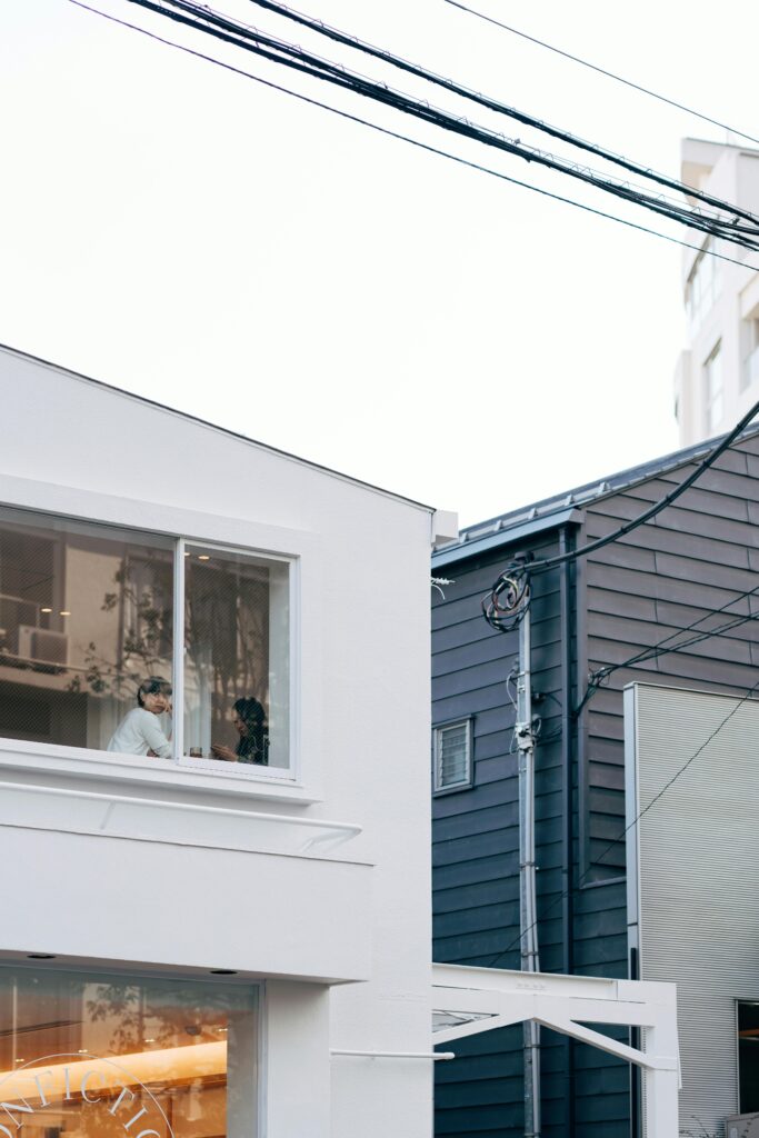 A quiet residential street scene in Tokyo with a modern white building, overhead wires, and two people visible through an upstairs window.