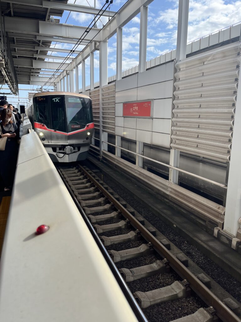 A Tsukuba Express train arriving at a station platform, with passengers waiting beside the tracks under a bright sky.