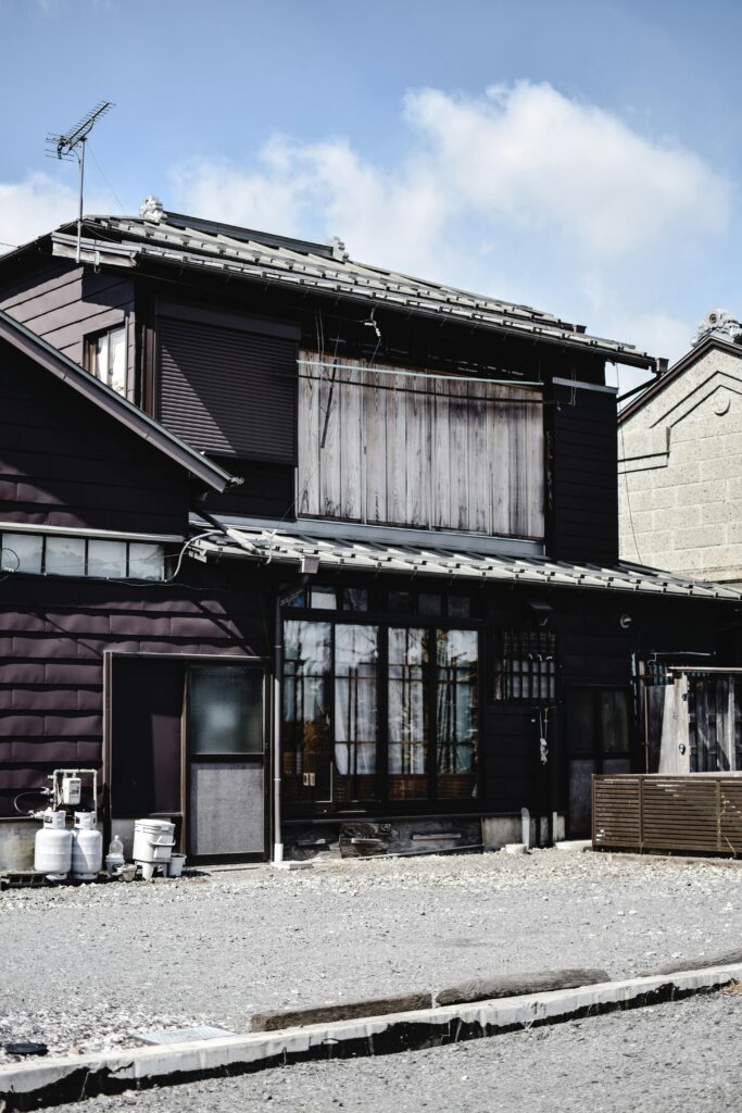 A traditional-style dark wooden house in Tsukuba with tiled roofing, sliding glass doors, and a simple gravel front area.