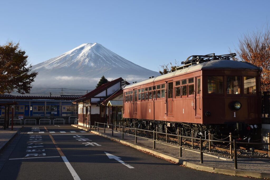 Vintage red train parked near a station with snow-capped Mount Fuji rising in the background under a clear blue sky.