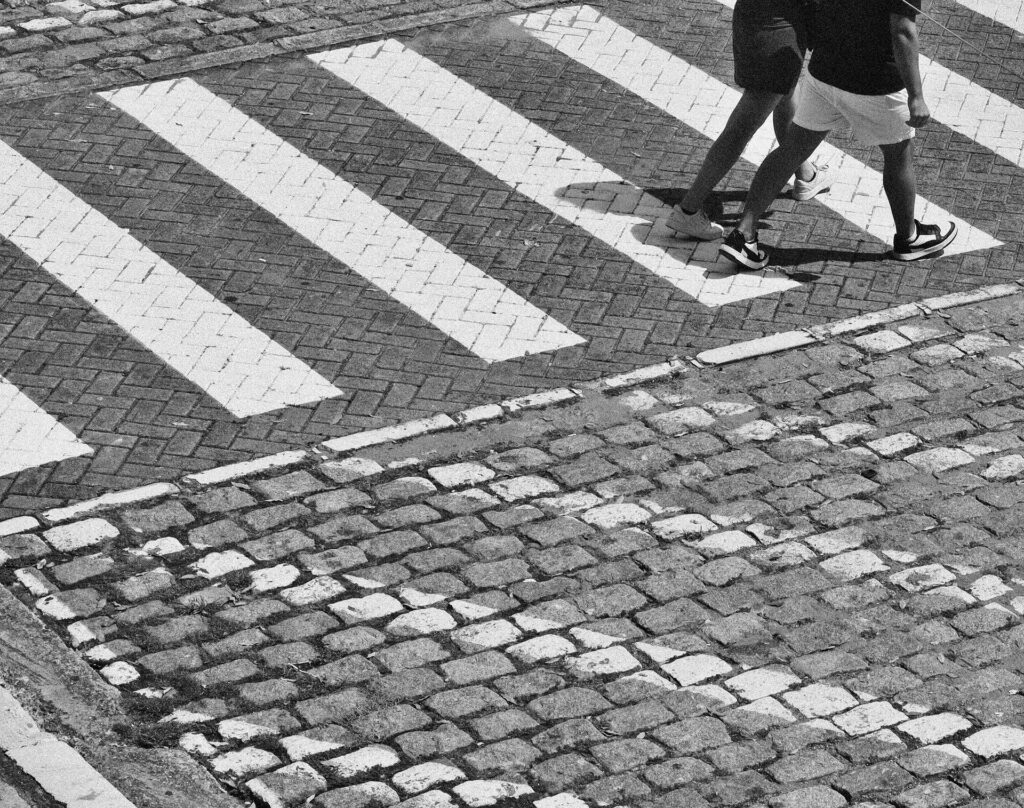 Black-and-white photo of two people walking across a zebra crossing on a cobblestone street.