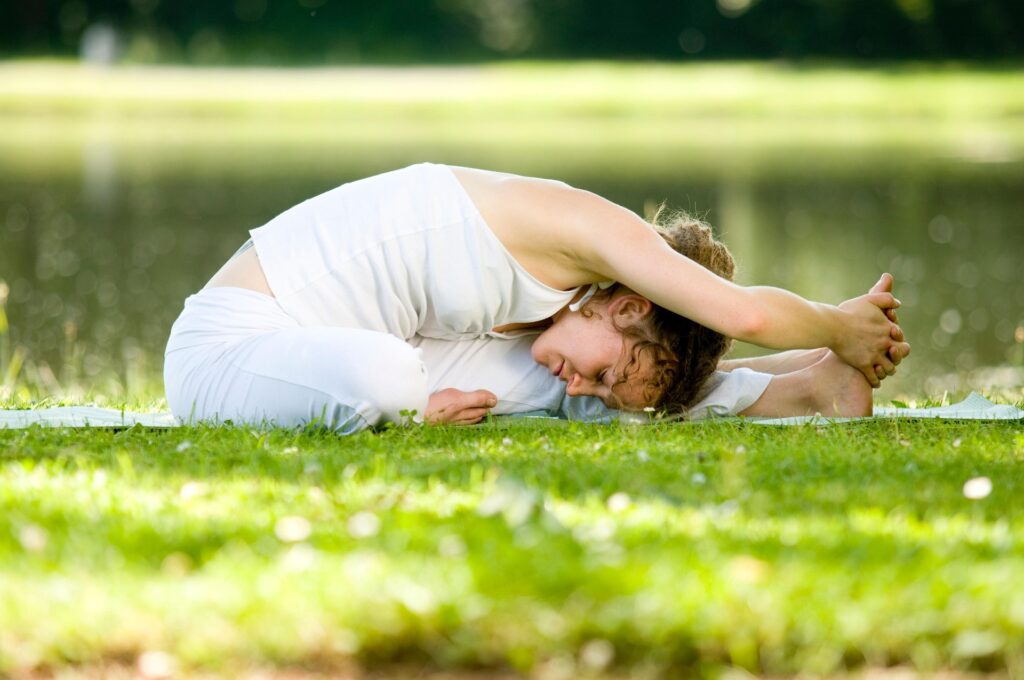 Person stretching on a yoga mat in a grassy park beside a calm lake.