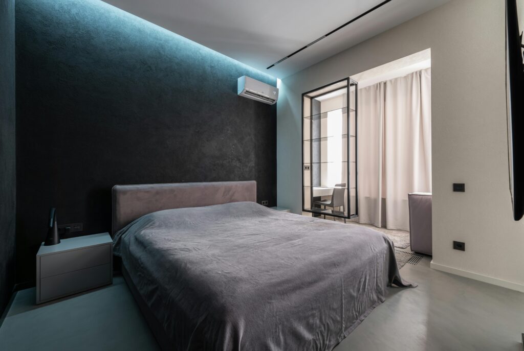 Modern bedroom with a wall-mounted air conditioner above the bed, dark accent walls, and soft ambient lighting.