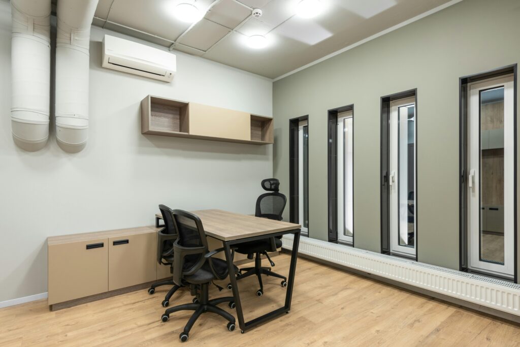 Small office room with a wall-mounted air conditioner, desk, rolling chairs, and tall narrow windows.