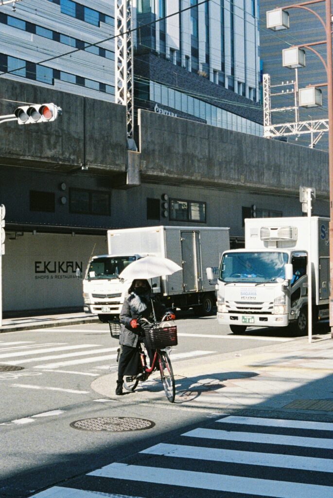 Woman riding a city bicycle with an umbrella at a Tokyo intersection while delivery trucks wait nearby