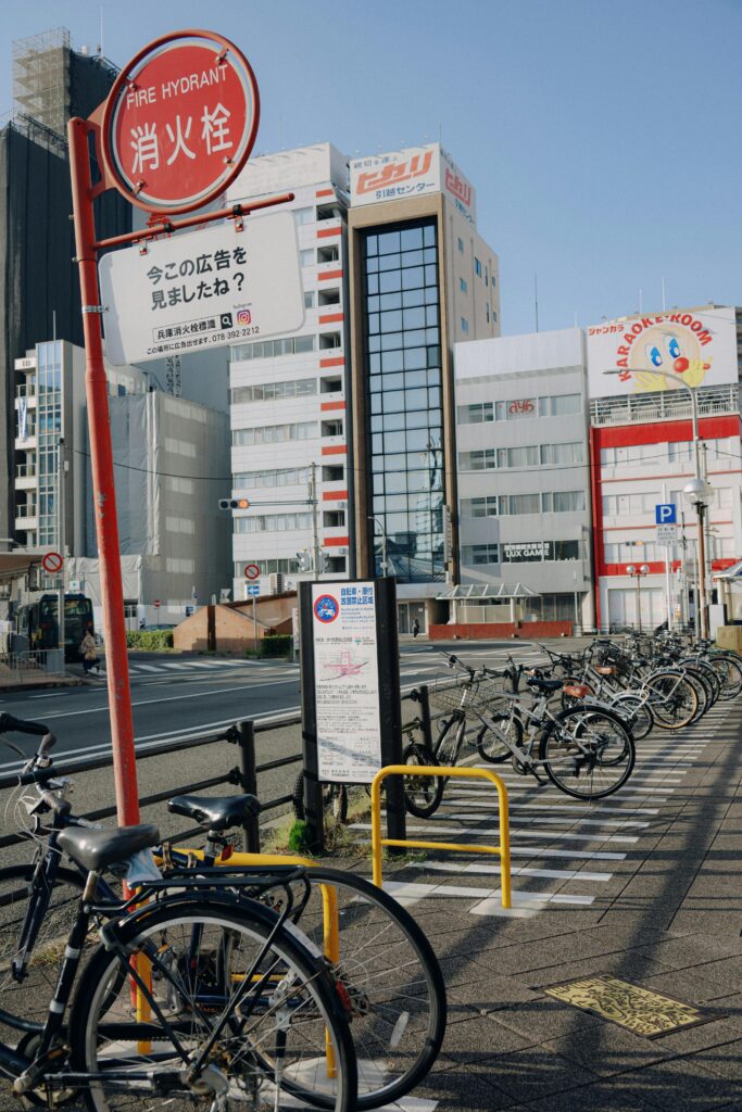 Bicycles lined up in an outdoor parking area beside a city street with warning signs and buildings in the background