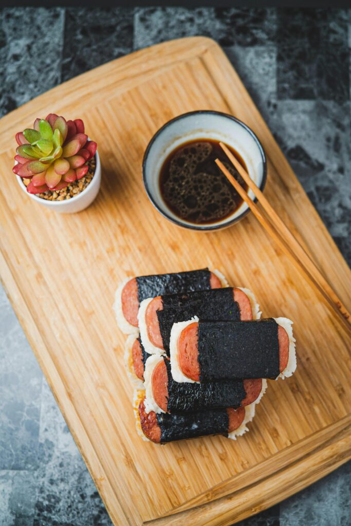 Spam musubi with grilled spam over rice wrapped in nori served on a wooden board with soy sauce