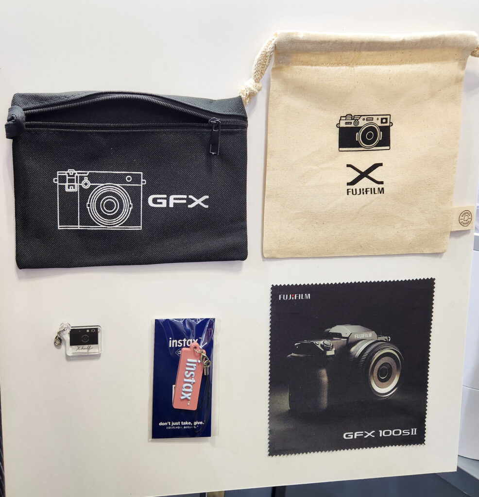 Fujifilm merchandise display with a GFX pouch, Fujifilm X drawstring bag, Instax keychain, branded tag, and GFX 100S II cleaning cloth.