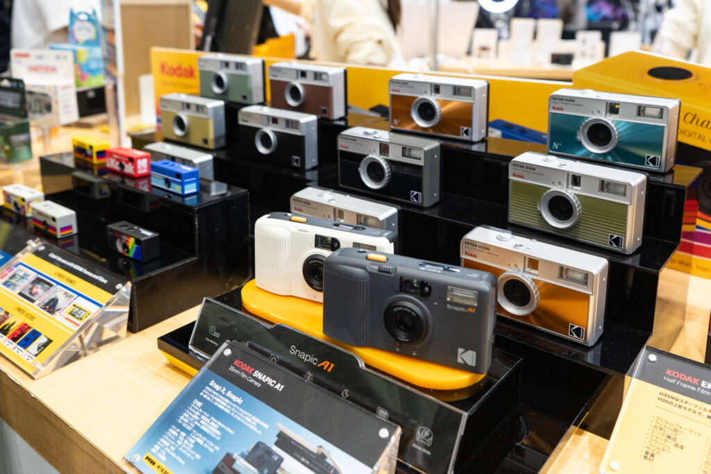 Kodak camera display featuring colorful compact and retro-style models, including Snapic and Ektar cameras, arranged on tiered stands.