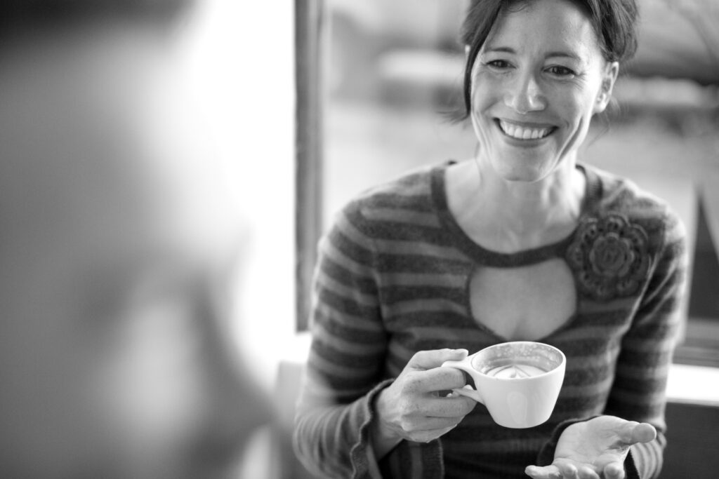 A smiling woman holding a cup of coffee while having a conversation in a relaxed indoor setting