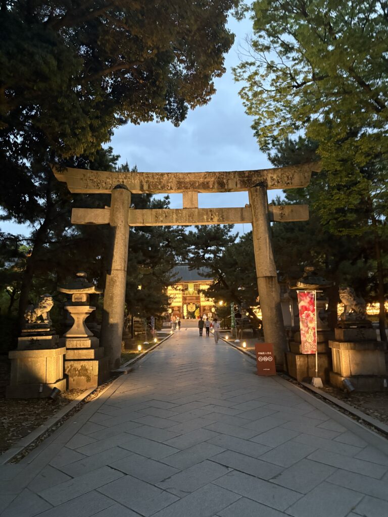Visitor standing on a bright red arched bridge surrounded by illuminated trees at Kitano Tenmangu Shrine during a nighttime Kyoto festival.
