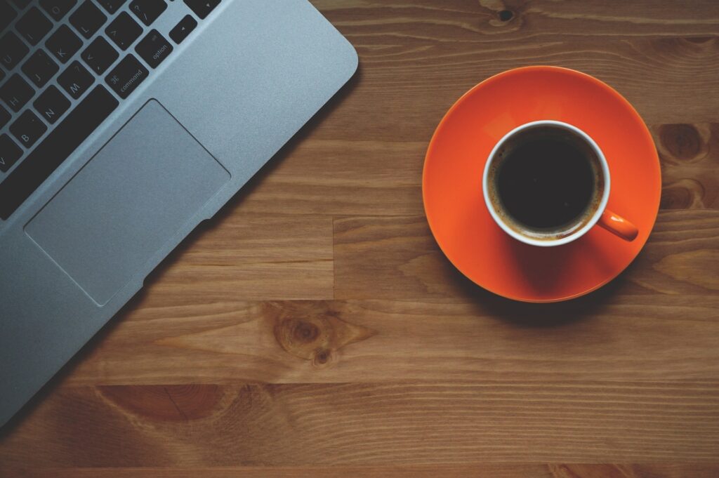 A laptop placed beside a cup of black coffee on a wooden desk, creating a cozy workspace scene.