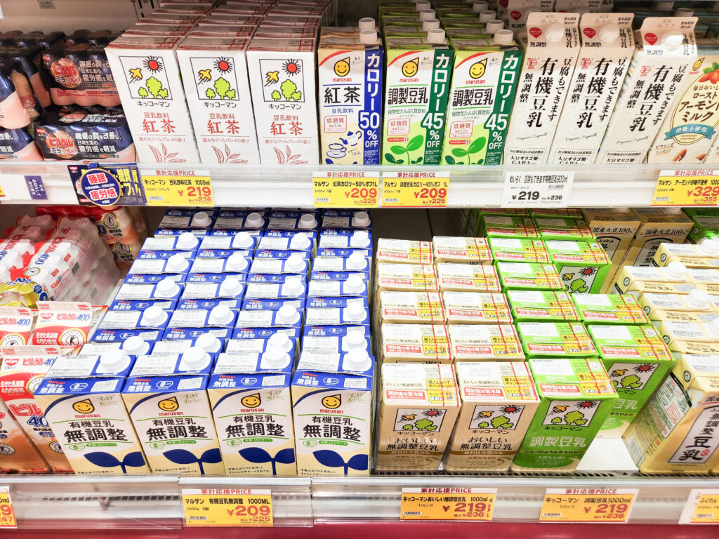 A refrigerated supermarket shelf in Japan displaying soy milk, almond milk, and dairy products with Japanese labels and pricing signs.