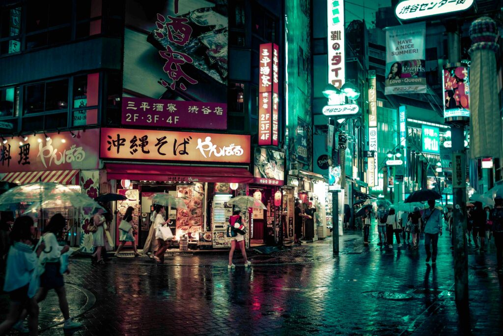 Rainy Tokyo street lined with restaurants and glowing signs as pedestrians walk with umbrellas on wet pavement at night.