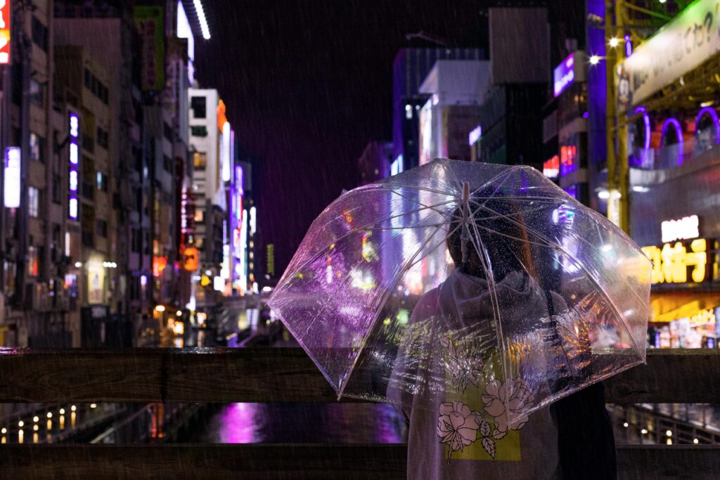 Person holding a transparent umbrella overlooking a rainy canal lined with neon lights in Osaka at night during wet weather in Japan.
