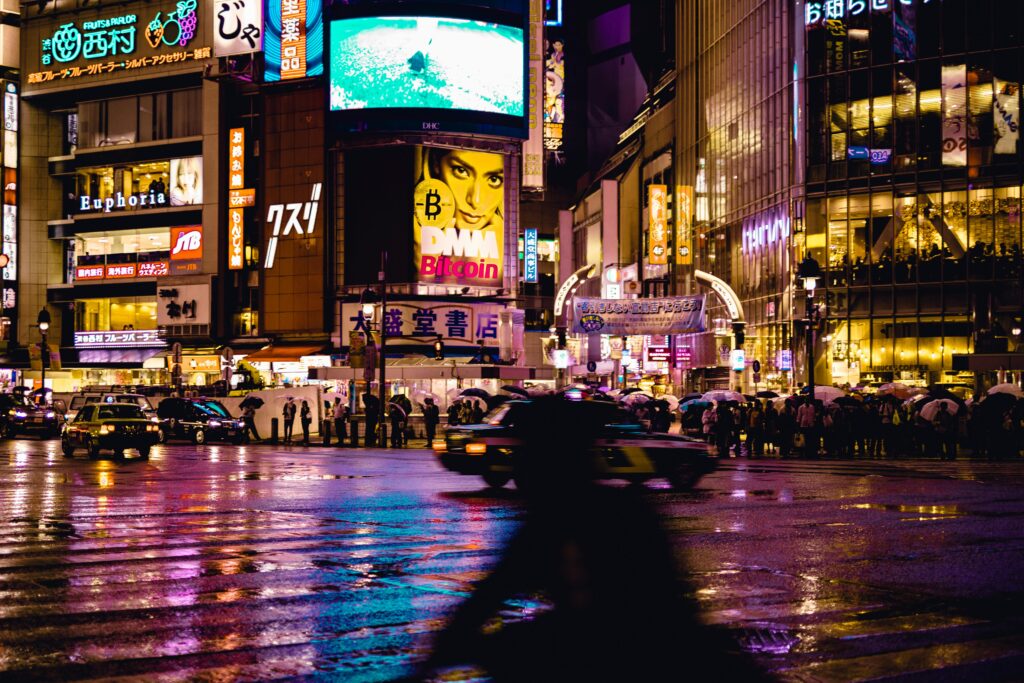 Rainy evening scene near Shibuya Crossing with glowing city lights reflecting on wet roads as pedestrians wait with umbrellas.