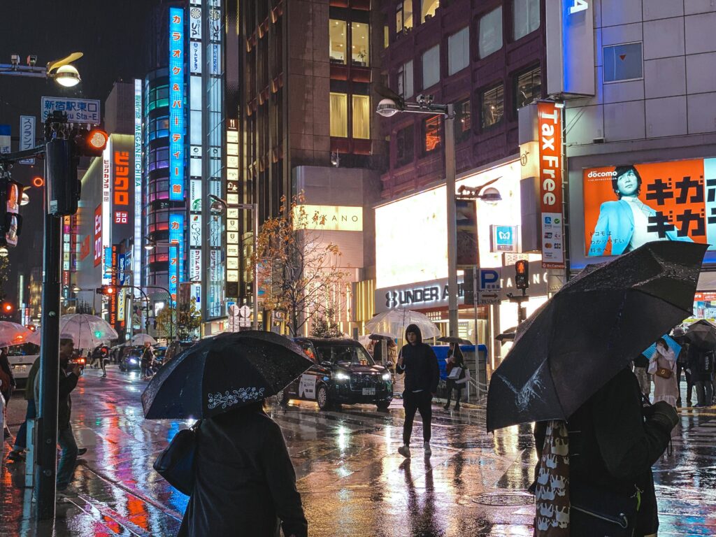 People walking with umbrellas along a rainy Tokyo street at night with reflections from bright city lights on wet pavement.