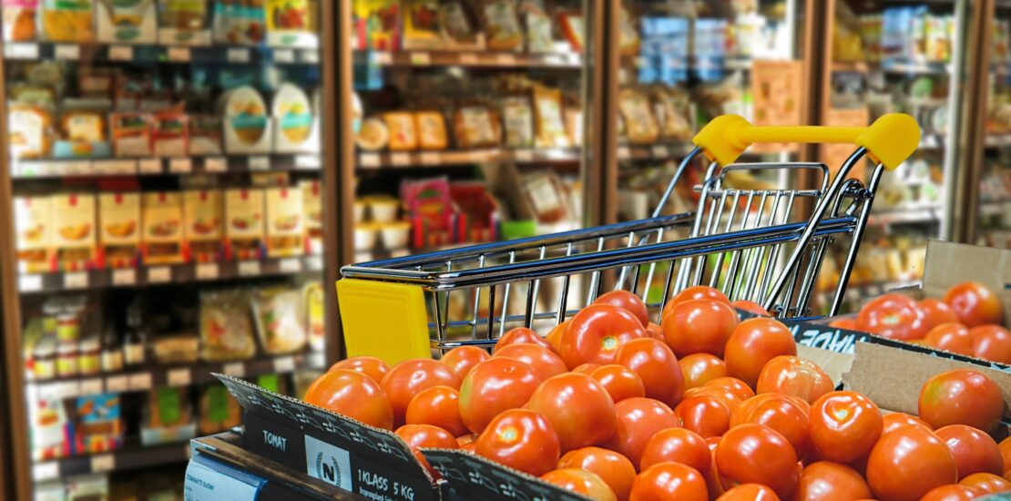 Shopping cart beside fresh tomatoes and refrigerated food shelves inside a modern supermarket grocery store.