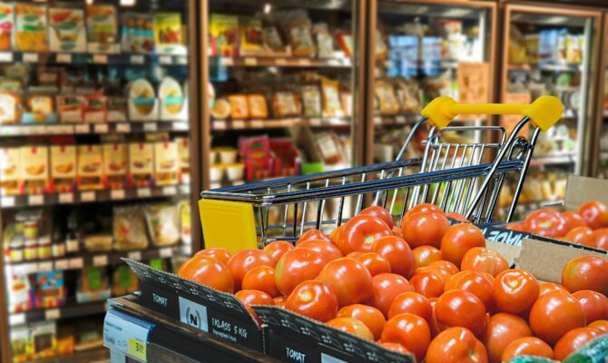 Shopping cart beside fresh tomatoes and refrigerated food shelves inside a modern supermarket grocery store.
