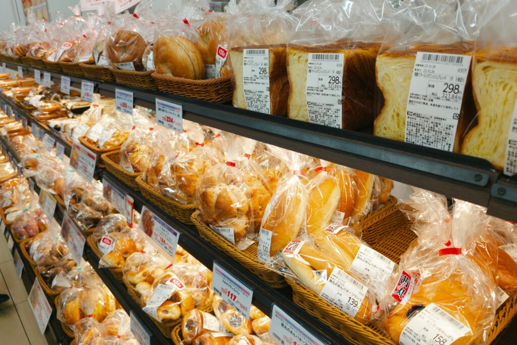 Packaged bread, pastries, and baked goods displayed on shelves inside a Japanese supermarket bakery section with visible price tags and baskets.
