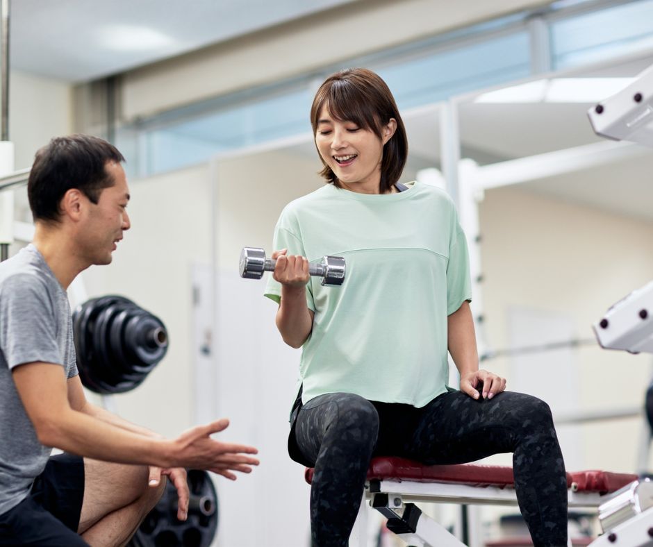 Woman lifting a dumbbell with guidance from a personal trainer inside a fitness gym in Japan.