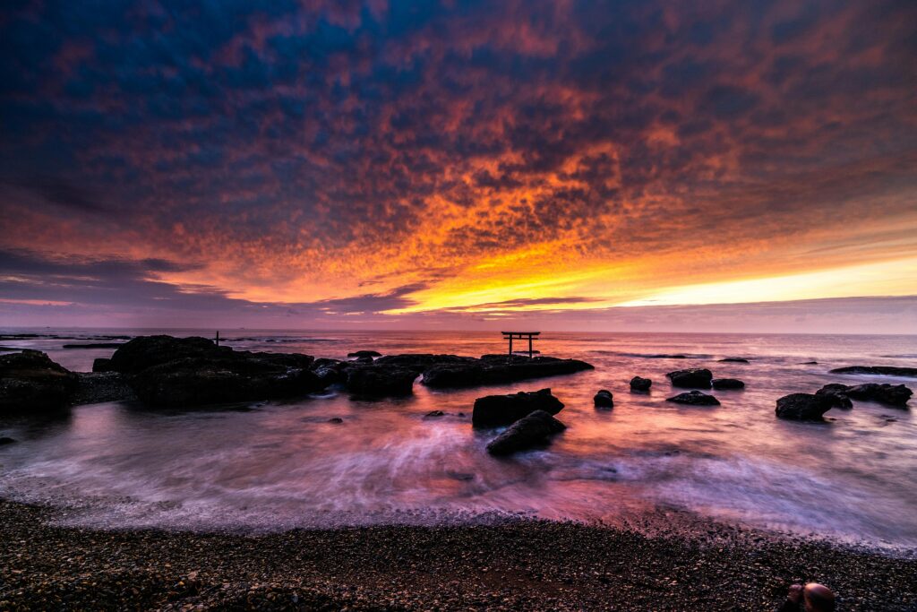Fiery orange and purple sunset over a rocky shoreline with gentle waves and a lone torii gate in the distance.