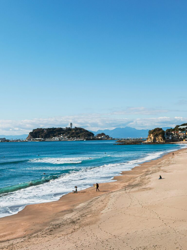 Wide sandy beach with rolling waves, Enoshima Island, and distant mountains under a clear blue sky.
