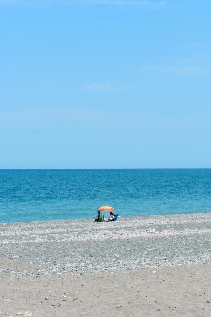 Two people relax under a colorful beach umbrella on a quiet pebble beach facing the calm blue sea.