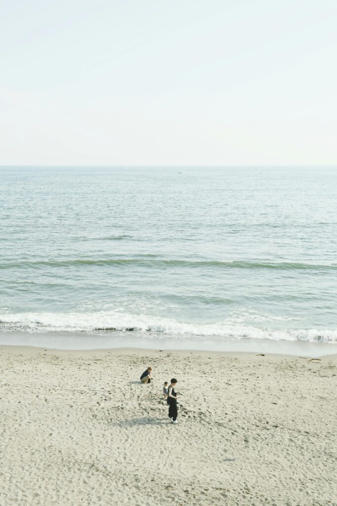 Two people walk across a wide sandy beach beside gentle waves under a bright, hazy sky.