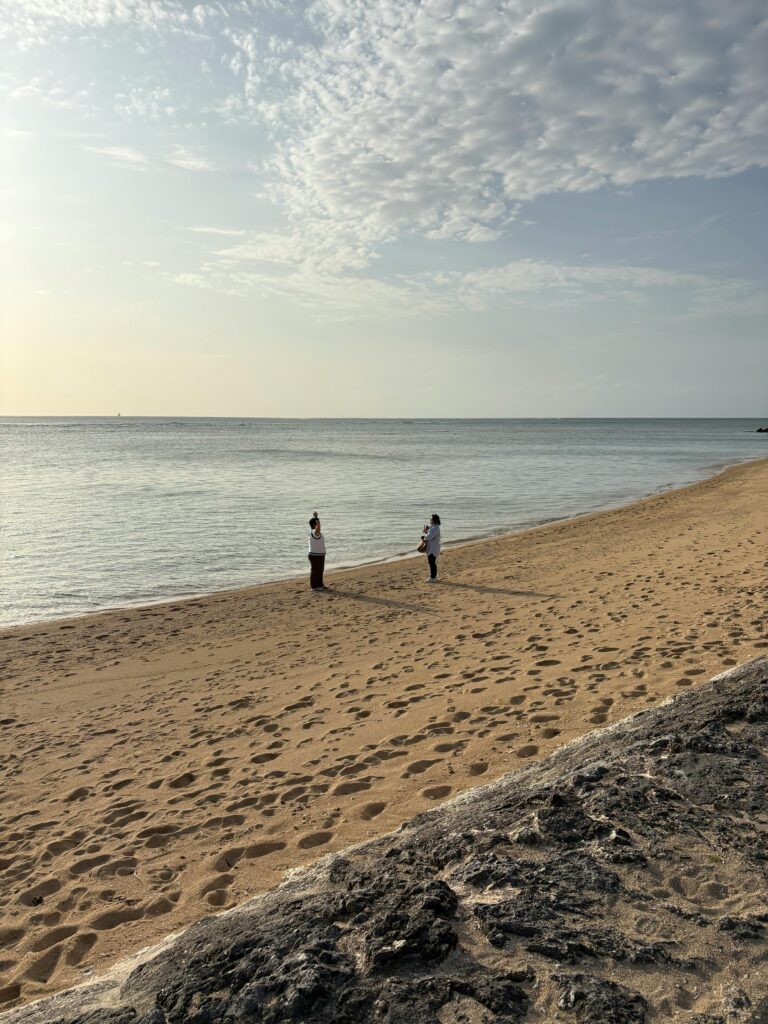 Two people stand on a peaceful sandy beach beside calm water under scattered clouds during golden hour.