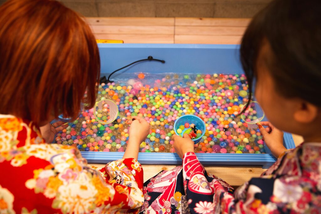 Visitors in colorful yukata scoop floating balls from a festival game booth during a Japanese neighborhood matsuri.