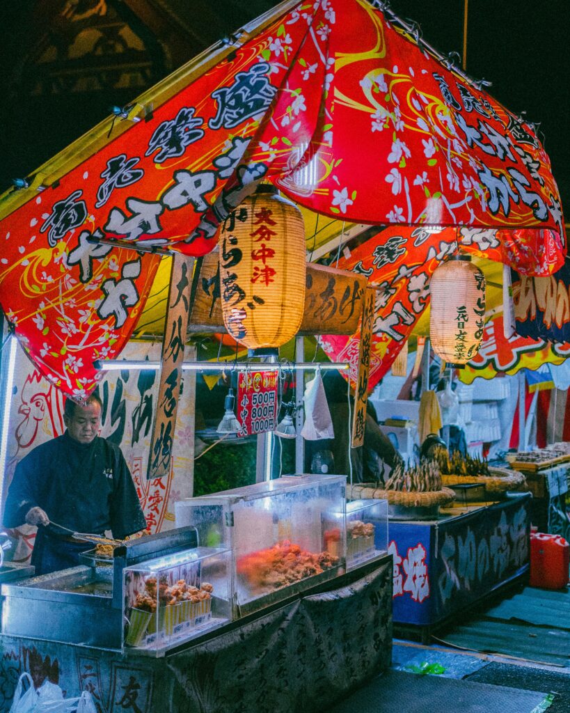 A food vendor prepares festival snacks at a brightly decorated yatai stall during a Japanese matsuri at night.