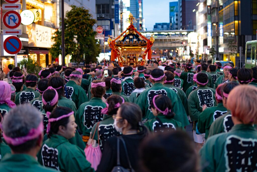 A large procession of participants in matching happi coats carries a mikoshi through the city during a neighborhood festival.