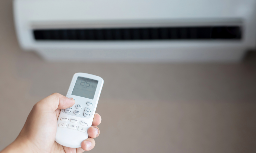 Hand using an air conditioner remote with a wall-mounted AC unit in the background.