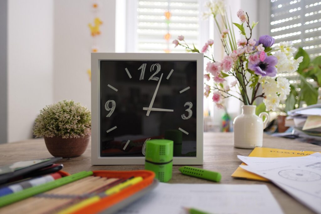 A study desk with a clock, stationery, notebooks, and flowers, representing time management and a productive homework routine.
