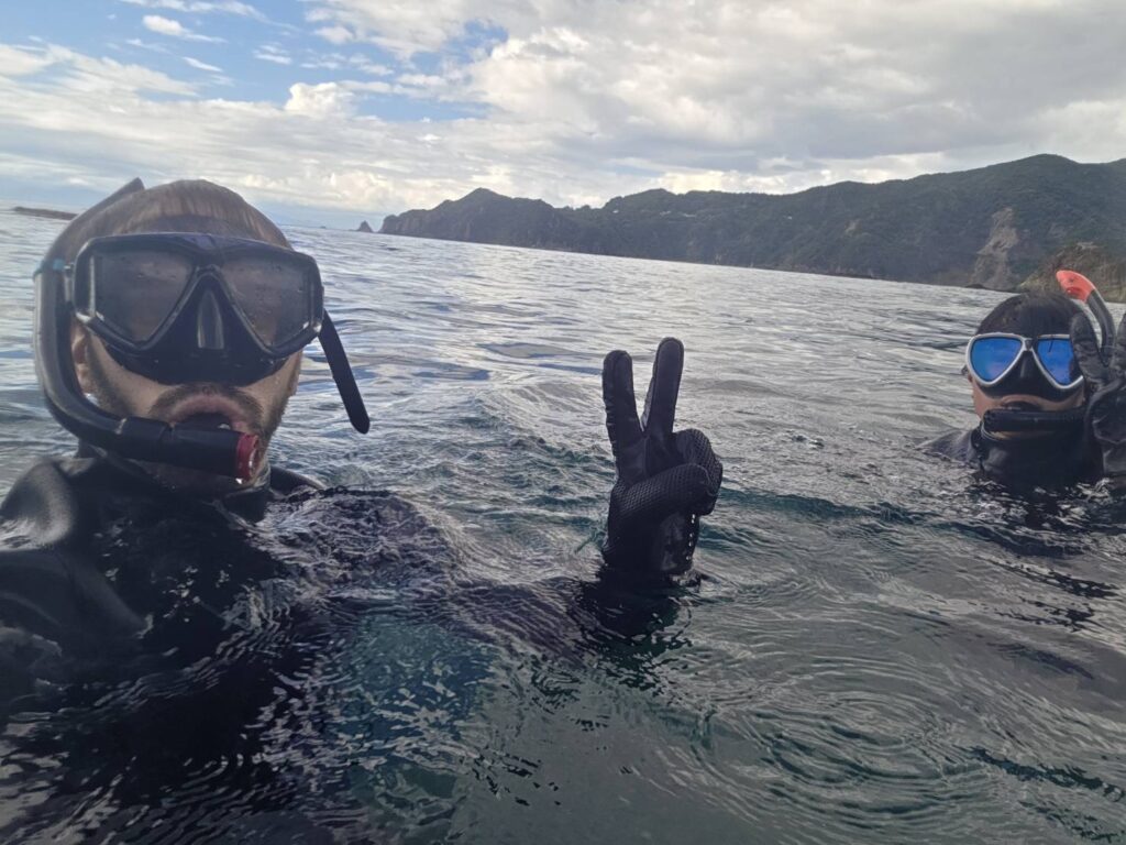 Two people snorkeling in the ocean at Koganezaki, Izu, with the rocky coastline and mountains in the background.