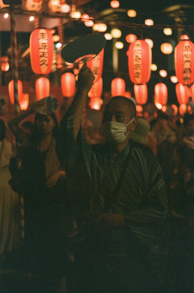 Person dancing with a fan beneath glowing red lanterns at a Japanese Obon festival.
