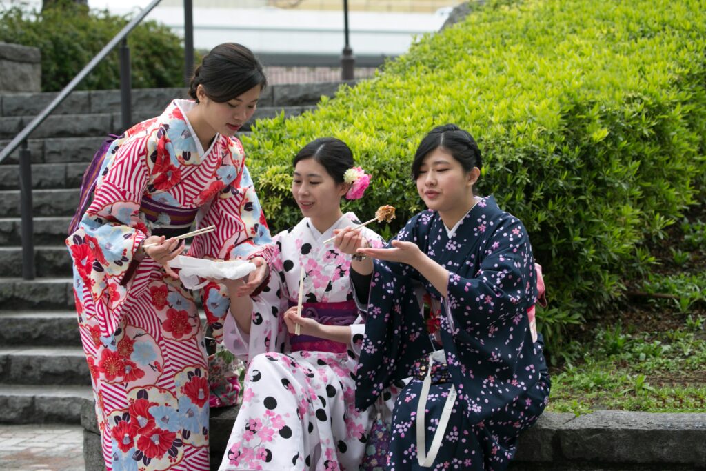 Three women wearing colorful yukata enjoy festival food together outdoors in Japan.